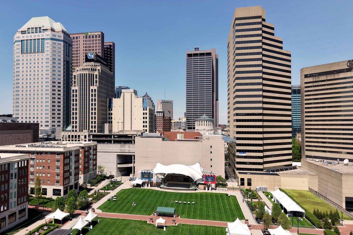 Downtown Columbus office towers rising above a riverfront park, with an open lawn and a white tensile-roofed amphitheater in the foreground.